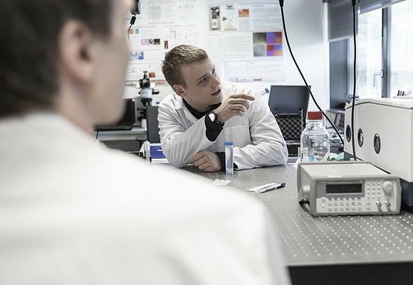 Ein Forscher in weißem Mantel erklärt einem anderen Forscher etwas. Im Hintergrund Laborgeräte ©Robert Josipovic